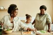 © Seventyfour - Medium shot of happy Black family of smiling mother, girl and grandmother laughing together while making healthy salad for dinner at beige kitchen