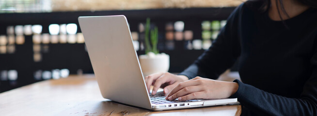 Woman hand typing laptop in cozy cafe. Woman in black long sleeves working online on computer at wooden table with natural lighting-banner image.