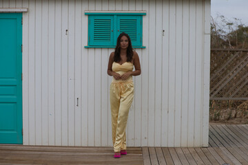 Wall Mural - Young and beautiful brunette woman poses self-confident in front of the white and turquoise wooden hut on a beach in Cadiz, andalusia, Spain. The woman makes different body postures like a model.