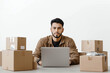 © alsu0112 - A young, handsome Hispanic man, an online store specialist, sits at a table with a laptop, surrounded by boxes of products, on a white background.