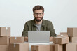 © alsu0112 - A bespectacled man of European appearance, an online store specialist, sits at a table with a laptop, surrounded by boxes of products, on a white background.