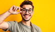 © Pixel Prophet - Waist up shot of cheerful brunet guy keeps hand on rim of spectacles wears casual t shirt and round spectacles being in good mood poses against vivid yellow background. Happy emotions concept isolat