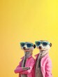 © jaykoppelman - Meerkat pair in pink jackets and blue sunglasses over yellow background.