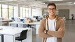 © lililia - A cheerful young Indian man stands in a contemporary office space, wearing a hoodie and glasses, showcasing a relaxed and confident demeanor while smiling warmly