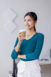 © David - Young successful employee business asian woman in blue shirt work hold takeaway delivery craft paper cup coffee to go stand at desk with laptop pc computer at office.