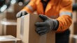 ©  Shomixer - An individual in an orange jacket, wearing gloves, carefully handles a cardboard box. The background features a warehouse setting, emphasizing logistics and manual labor.