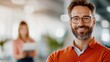 © LifeMedia - A bearded man with glasses smiles confidently while standing in a modern office environment, conveying a sense of achievement and professional satisfaction.