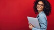 © farhan - Happy female student with curly hair and glasses, holding a laptop, on a cherry red background, copy space