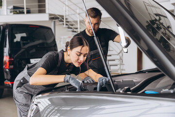 Trying to identify the source of the problem. Two mechanics man and woman working together at open hood on a car in an auto repair shop.