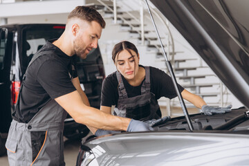 Trying to identify the source of the problem. Two mechanics man and woman working together at open hood on a car in an auto repair shop.