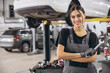 © anatoliycherkas - Smiling young woman mechanic standing beneath lifted car, holding tools and looking at camera in car service garage