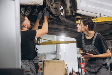 Young expert female inspects repair checklist with automotive mechanic worker partner, quality suspension technician team at fix garage. Vehicle maintenance service works industry occupation job.