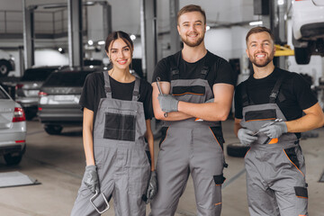 Wall Mural - Group of three car service technician men and woman talking at workplace, people working together at vehicle repair garage service shop, check and repair customer car at automobile service center.