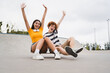 © InsideCreativeHouse - Couple of happy friends teenagers boy girl classmates high school pupils students in colorful t-shirts holding hands up celebration have fun looking at camera in skate ramp park sitting on skateboard
