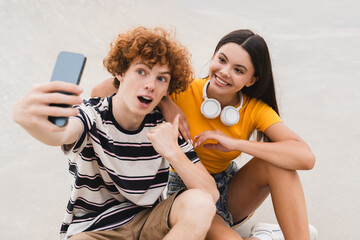 Wall Mural - Couple of teenagers classmates students high school pupils friends boy girl bloggers in bright colorful casual holding smart mobile phone taking selfie shot sitting on skate park ramp after lessons