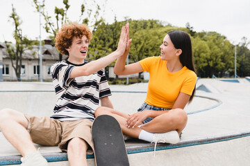 Couple of happy friends teenagers boy girl classmates high school pupils students giving high five celebration have fun looking to each others sitting in skate ramp park riding on skateboard