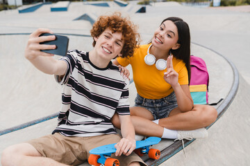 Wall Mural - Couple of teenagers classmates students high school pupils friends boy girl bloggers in casual with skateboards bags taking selfie shot holding smart mobile phone having fun sitting on skate park ramp