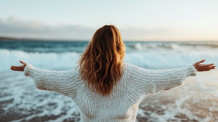 A woman wearing a warm sweater is standing on the beach with her back to the camera, spreading her arms wide open towards the sea, capturing the sense of freedom and relaxation.