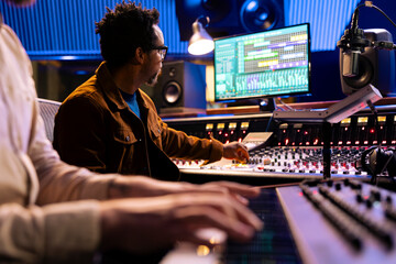 Wall Mural - African american audio expert records artist playing piano in studio, using mixing console and digital editing software on pc. Producer composing new songs with musician on electronic keyboard.