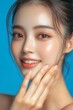 © Eyestetix Studio - Skin care. Woman with beauty face touching healthy facial skin portrait. Beautiful smiling asian girl model with natural makeup touching glowing hydrated skin on blue background