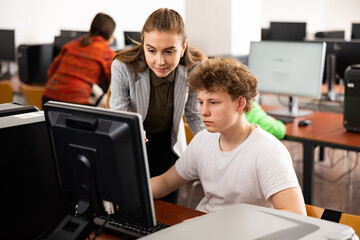 Young female teacher helping focused teenage student studying for exam in college library computer lab..