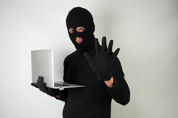 Wall Mural - Young Adult black masked robber wearing gloves is holding silver colored laptop for hacking, scamming and phishing.