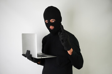 Wall Mural - Young Adult black masked robber wearing gloves is holding silver colored laptop for hacking, scamming and phishing.