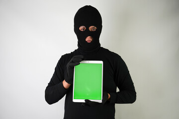 Wall Mural - Young Adult black masked robber is holding digital tablet with vertical blank green screen. Concept for hacking, scamming, phishing.