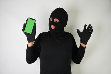 Wall Mural - Young Adult black masked robber is holding smartphone with blank green screen. Concept for hacking, scamming, phishing.