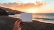 © Nata - A person is holding a white square piece of paper on a beach. The paper is blank, and the person is looking at the ocean. Concept of calmness and serenity