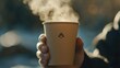 © DREAMERLYN STUDIO - Close-up of a hand holding a paper cup with a steaming hot coffee, featuring a stylish logo and sustainable packaging design.