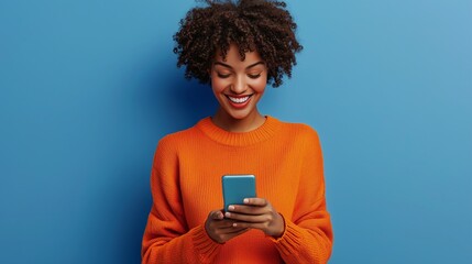 Photo of cheerful delighted african american woman types sms on modern cell phone device enjoys good internet connection dressed in orange jumper focused aside isolated on blue studio wall.