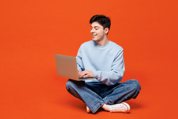 Wall Mural - Full body young smart Caucasian IT man he wears blue sweatshirt casual clothes sits hold use work on laptop pc computer isolated on plain red orange color background studio portrait Lifestyle concept