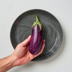 Wall Mural - A hand holds a fresh, purple eggplant on a gray plate, white background.