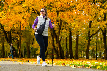 Wall Mural - Smiling middle-aged blonde woman dressed sporty walking with poles on background of beautiful autumnal trees. Front view. Autumnal training of Nordic walking.