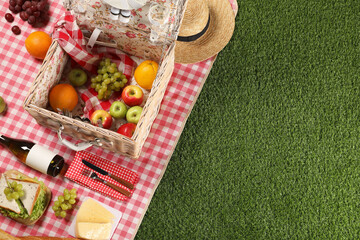 Wall Mural - Picnic basket with tableware, different snacks, wine and hat on green grass outdoors, above view. Space for text