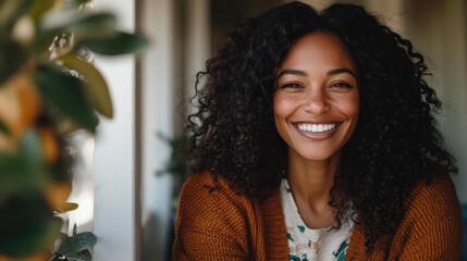 A woman with curly hair, smiling brightly while sitting in natural lighting indoors, wearing a cozy orange sweater, capturing a moment of happiness and relaxation in a warm environment.