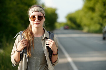 young female backpacker in sunglasses, headband and casual attire looking at camera while standing b