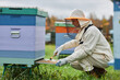 © Seventyfour - Side view of serious mature female beekeeper in protective bee suit cleaning wooden colorful beehive scraping dirt off bottom board at apiary farm