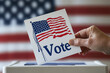 © abderahim - man is voting in an USA election by putting a white piece of paper into a box. USA flag on background.