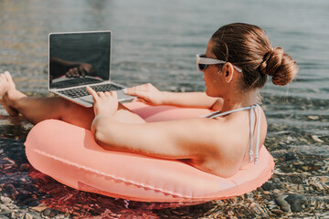 Wall Mural - A woman is sitting on a pink inflatable raft in the water, using a laptop. Concept of relaxation and leisure, as the woman is enjoying her time by the water while working on her laptop.