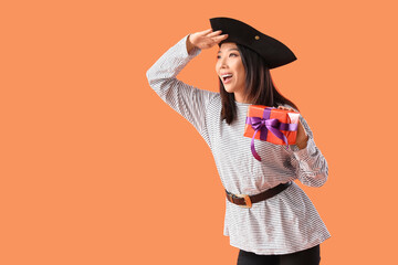 Poster - Young Asian woman dressed as pirate with Halloween gift on orange background