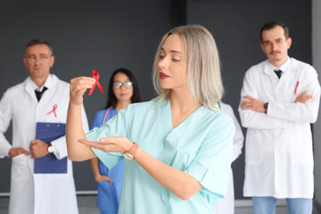 Wall Mural - Young female medic with red ribbon and group of doctors in clinic. World AIDS Day concept