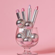 © sunaiart - A silver ballpoint pen and lipstick are set inside an ice cream glass, surrounded by disco balls on a pink background, creating a pastel, whimsical aesthetic.