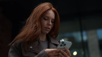 Low-angle view of cheerful cute young woman wearing fashion leather jacket using typing mobile phone smiling looking to screen, happily using smartphone app walking in city on cloudy day.
