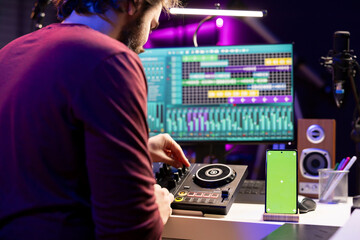 Wall Mural - Music producer uses controls and knobs on mixing console in home studio, having a smartphone with greenscreen display. Artist learning to edit tunes and add sound effects with daw software.