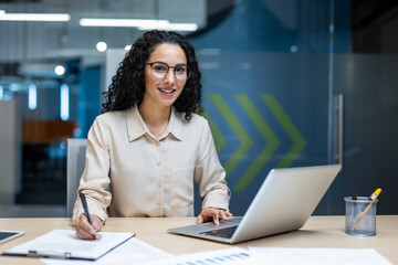 Wall Mural - Confident businesswoman working at office desk, using laptop and taking notes. Professional setting with focus on productivity and success.