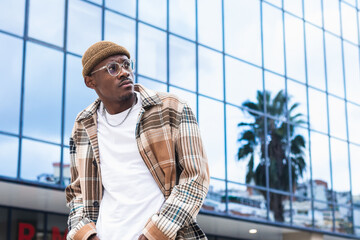 Trendy black guy walking on street. Young African American male in stylish checkered shirt looking away and walking on city street outside modern glass building