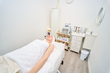 A woman is laying on a table in a spa. The room is clean and well-lit. The woman is wearing a white towel and has a bottle of lotion on the table