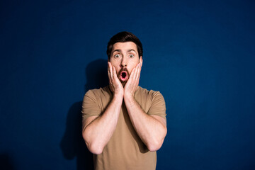 Wall Mural - Photo of speechless man with beard dressed beige t-shirt astonished staring hold hands on cheeks isolated on dark blue color background